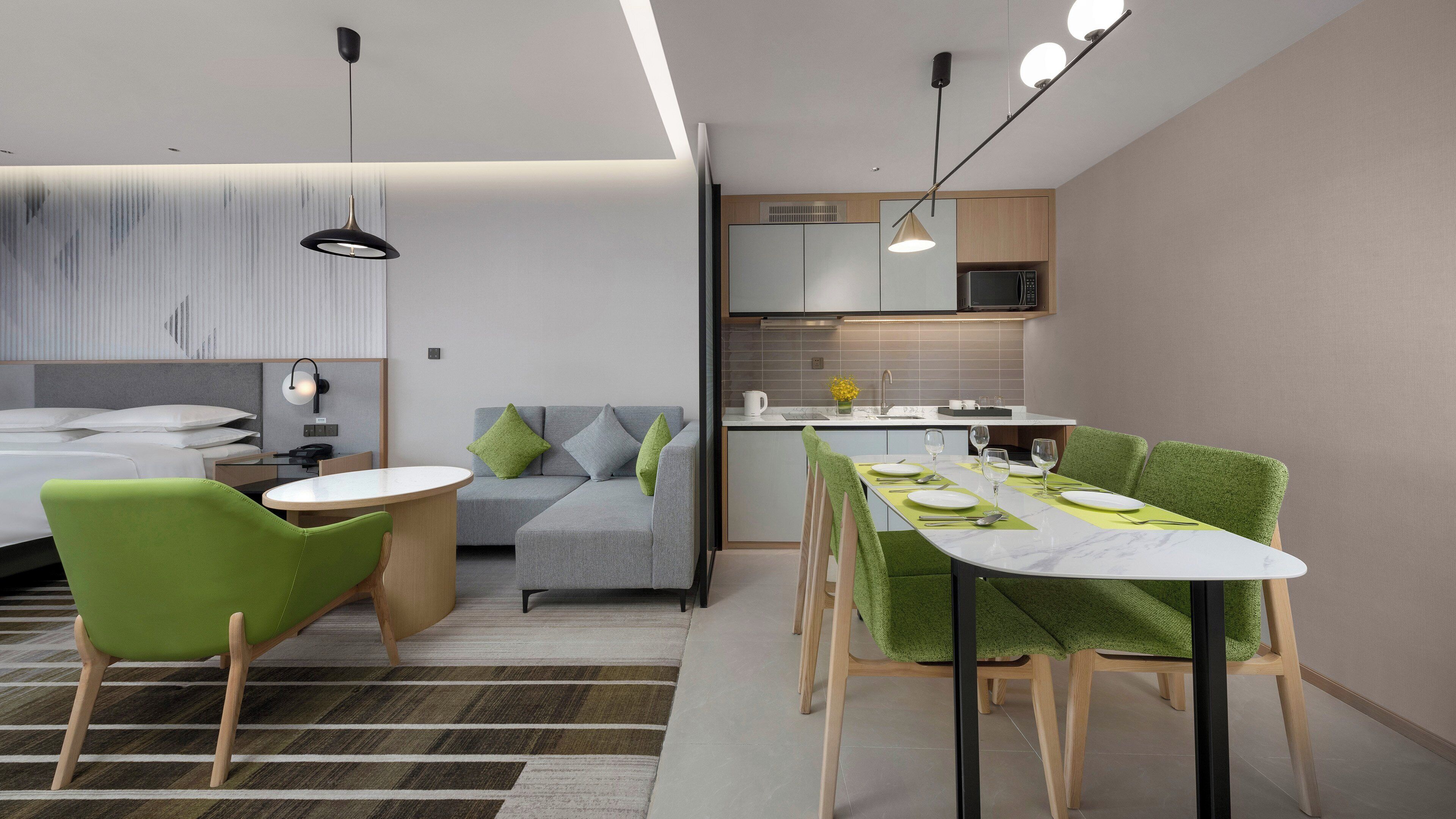 A modern hotel room with a bed, a sofa, a dining table, and a kitchen area.