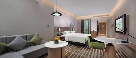 Une chambre d’hôtel moderne avec un canapé gris, un lit avec du linge de maison blanc, une petite table ronde ornée d’un vase de fleurs et un bureau avec une chaise.