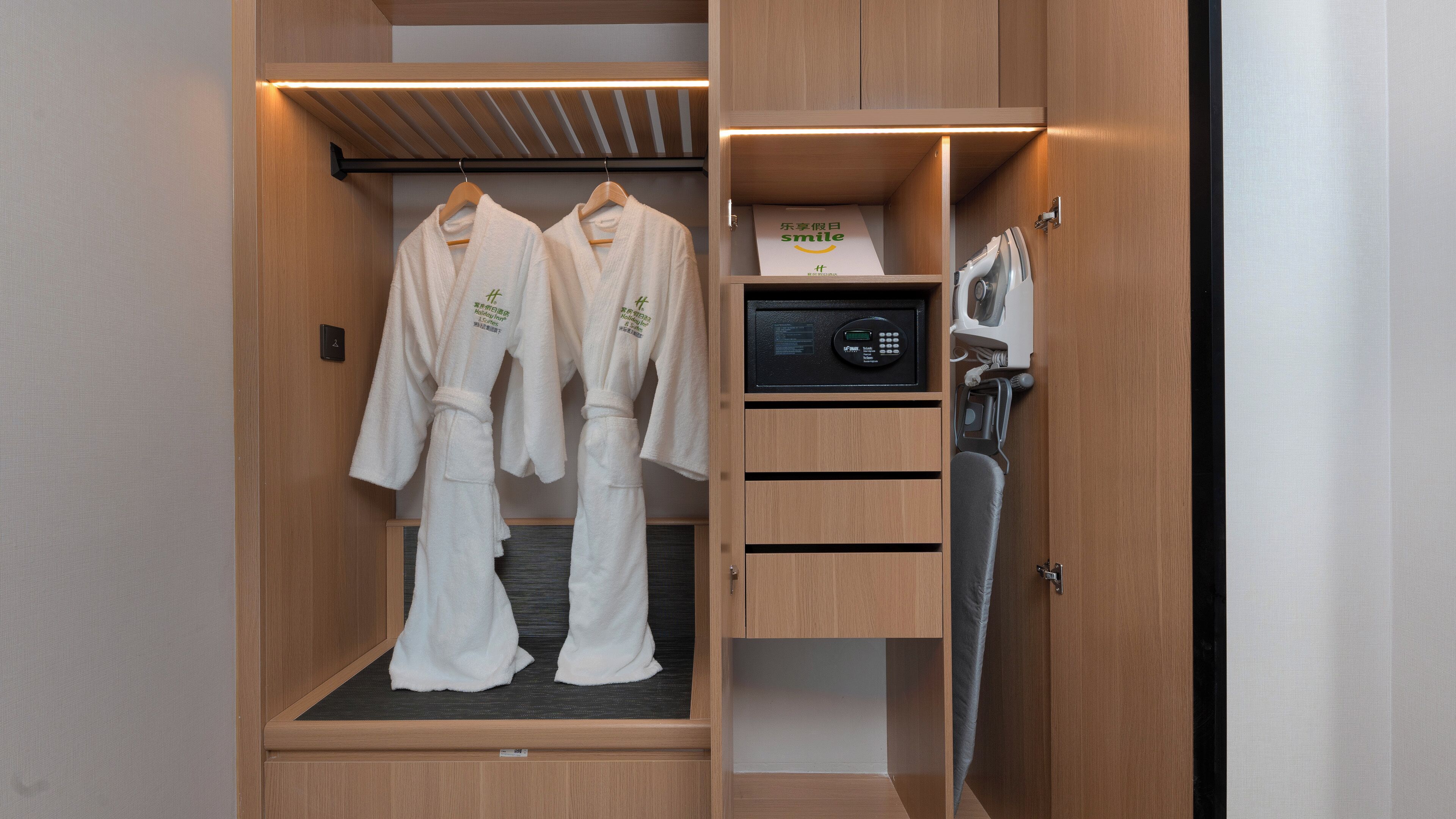 A hotel room with a wooden wardrobe containing two white robes, a microwave, and a small shelf with a book.