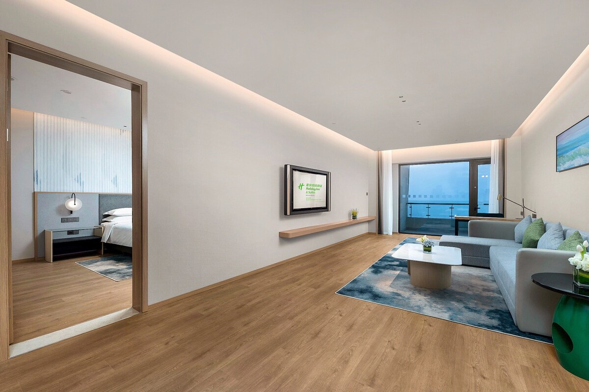 Habitación de hotel moderna con suelo de madera, un sofá, una mesa de centro y un gran ventanal con vistas.