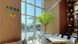 Een hal met een houten wand, een groot raam, een boom en een zithoek met tafels en stoelen.