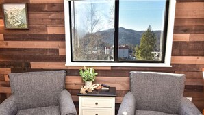 Una habitación con dos sillones grises tapizados, una pequeña mesa blanca con una planta y un ventanal con vistas a montañas y edificios.