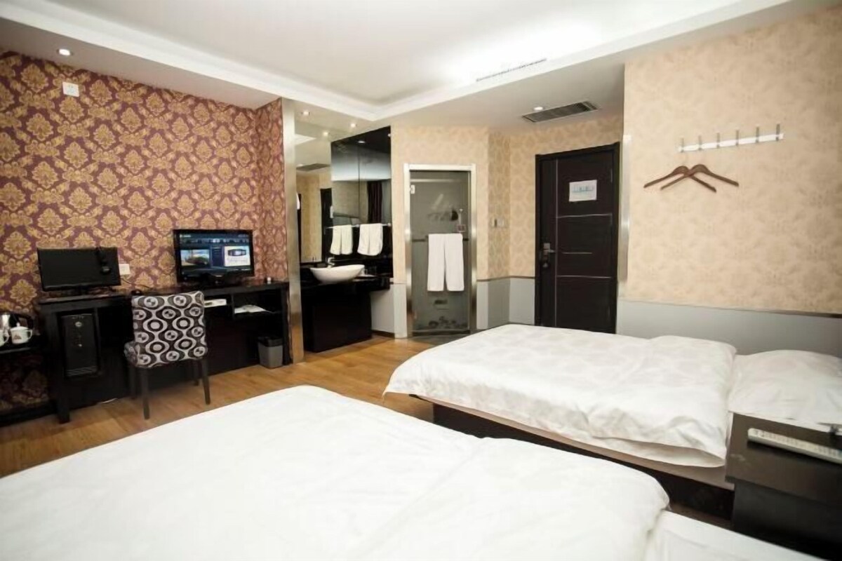 A hotel room with two beds, a desk with a computer, a chair, a TV, and a bathroom with a mirror and towel rack.