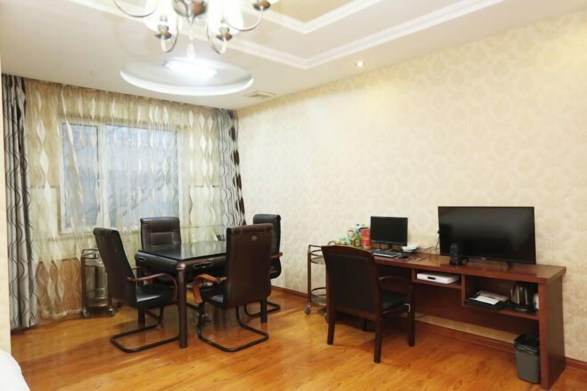 A room with a wooden floor, a dining table with chairs, a TV on a wooden stand, and a chandelier.