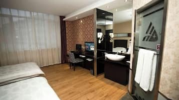 A hotel room with a bed, a desk with a computer, a bathroom with a sink and mirror, and a large window with curtains.
