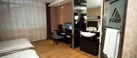 A hotel room with a bed, a desk with a computer, a bathroom with a sink and mirror, and a large window with curtains.