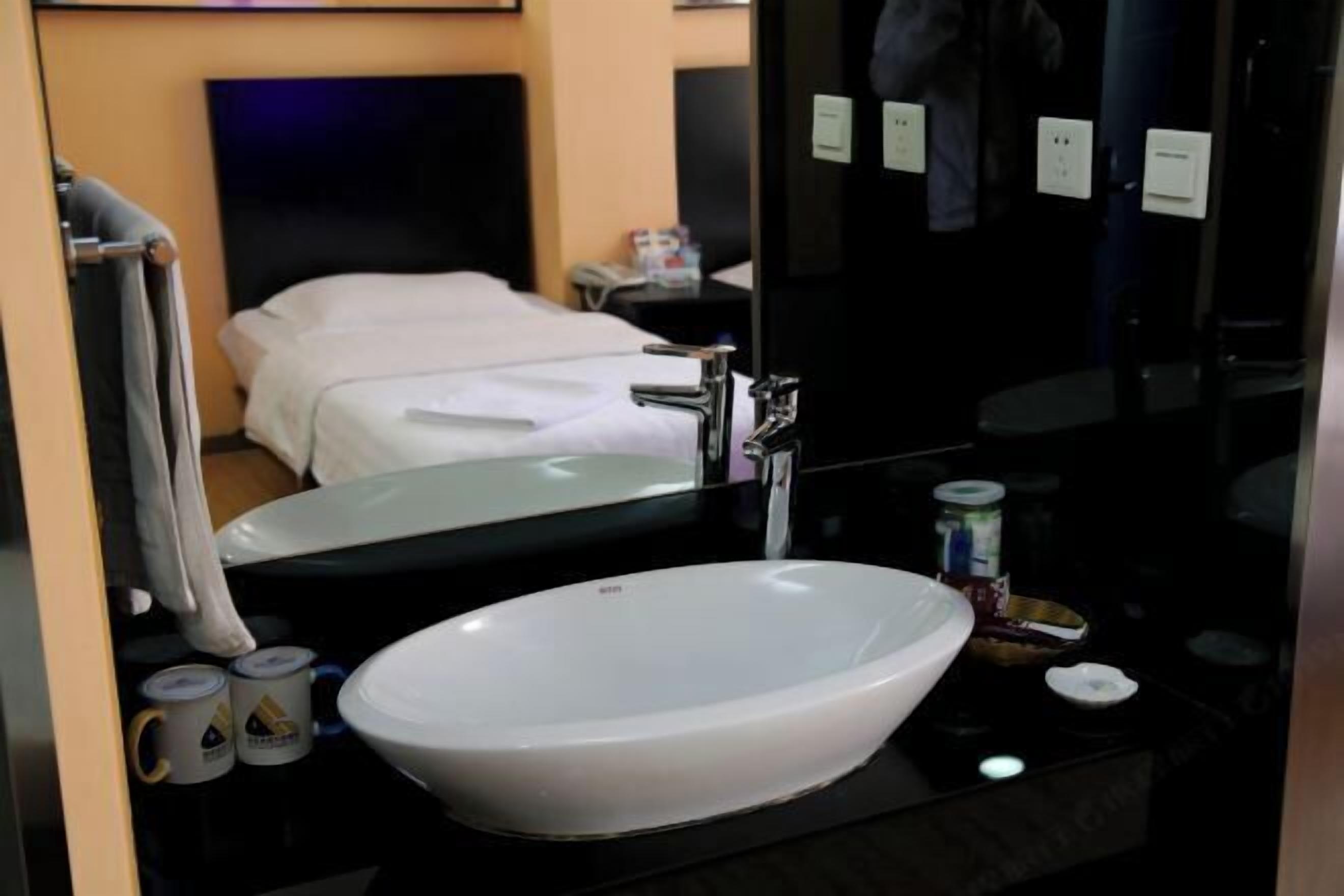 A modern hotel bathroom with a large oval sink, a mirror, and a bed in the background.