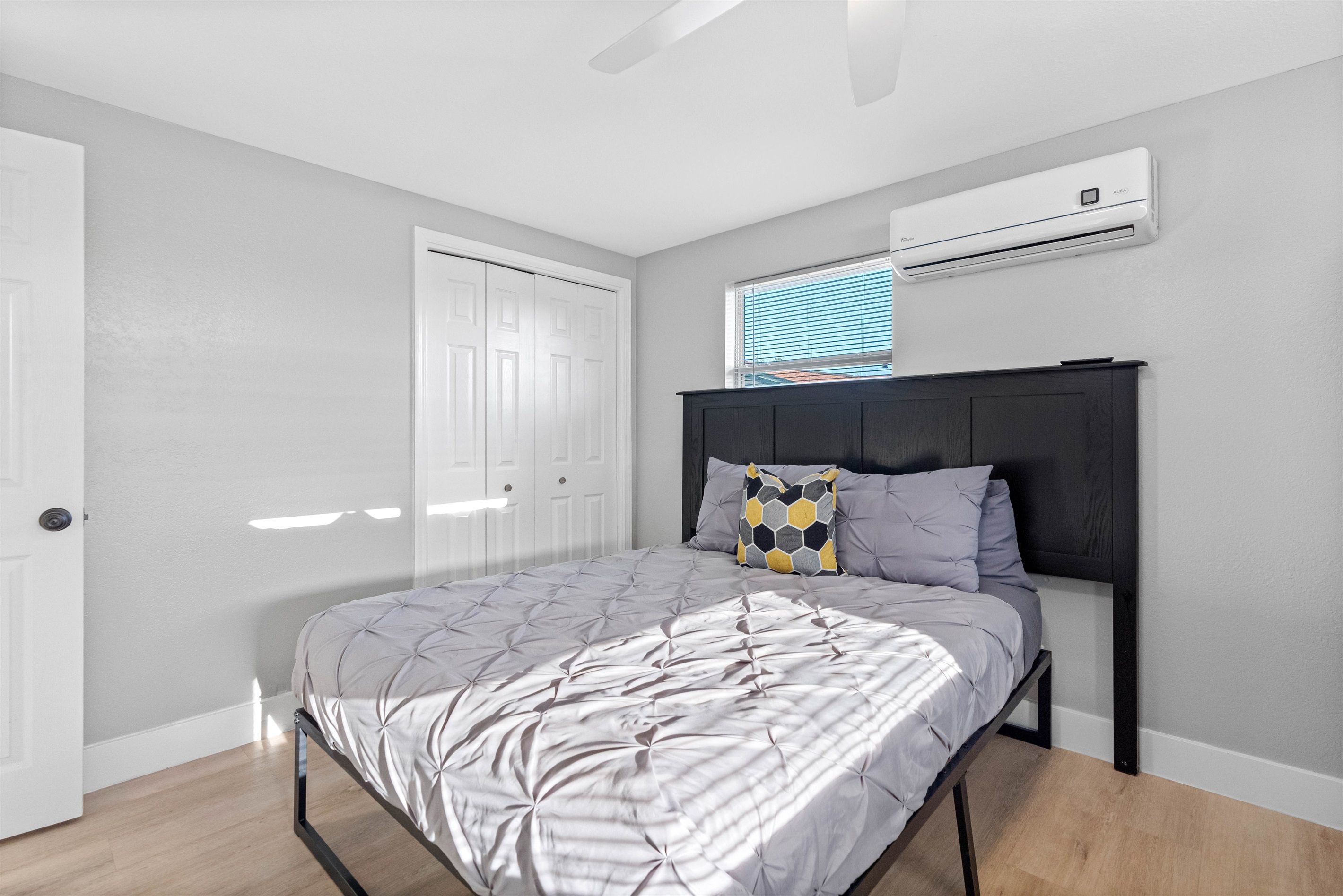 A bedroom with a bed, an air conditioning unit, a window with blinds, and a door.