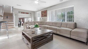 A modern living room with a sectional sofa, a wooden coffee table, a dining area with a bar stool, and a staircase leading to another level. - Luxury Heated Salt Pool House in Perfect Location (Tampa)