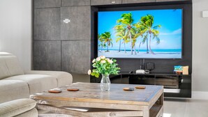 A modern living room with a flat-screen TV mounted on a dark wall, a wooden coffee table with a vase of flowers, and a beige sofa. - Luxury Heated Salt Pool House in Perfect Location (Tampa)