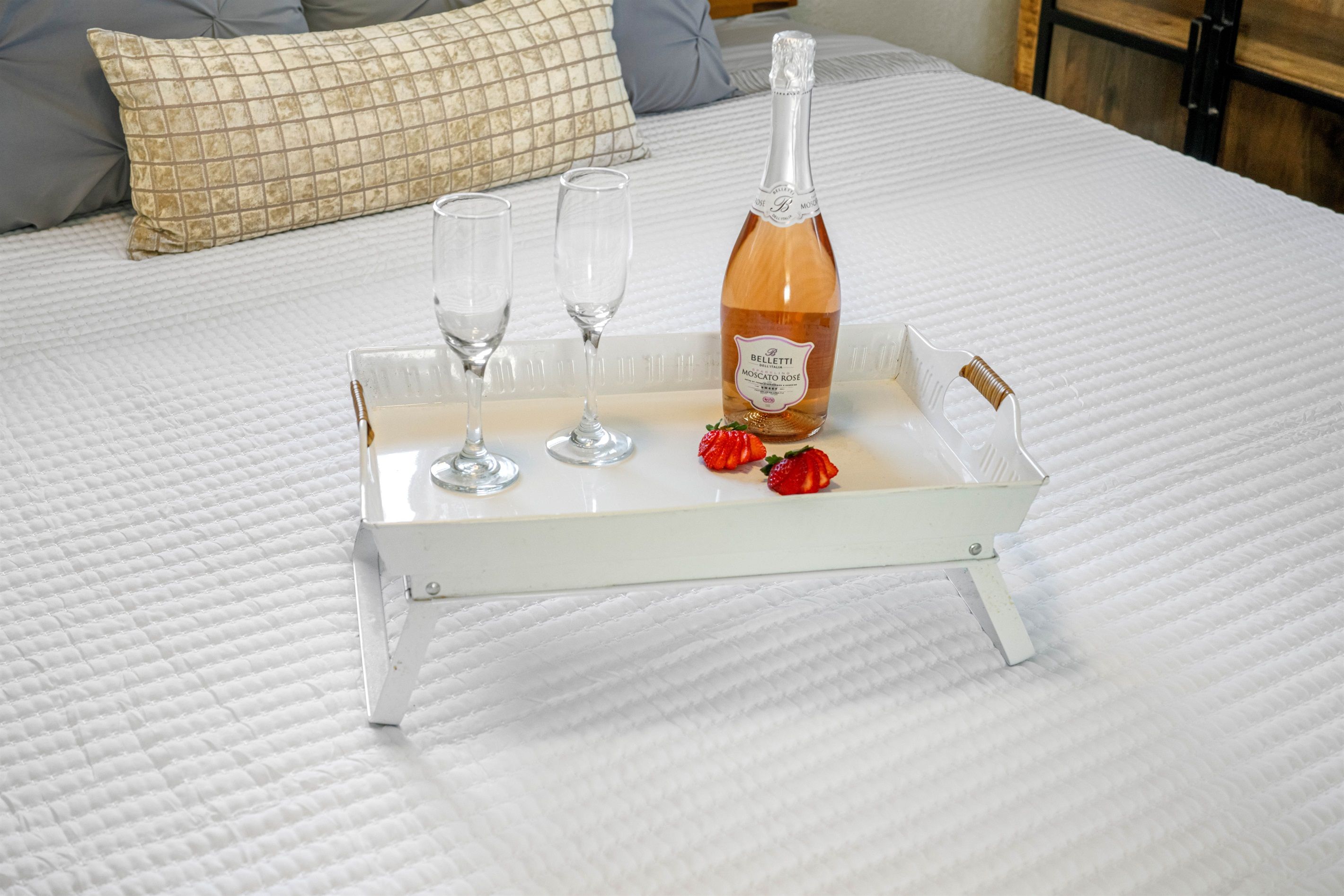 A bed with a white quilted bedspread, a white tray with a bottle of champagne, two glasses, and a few strawberries.