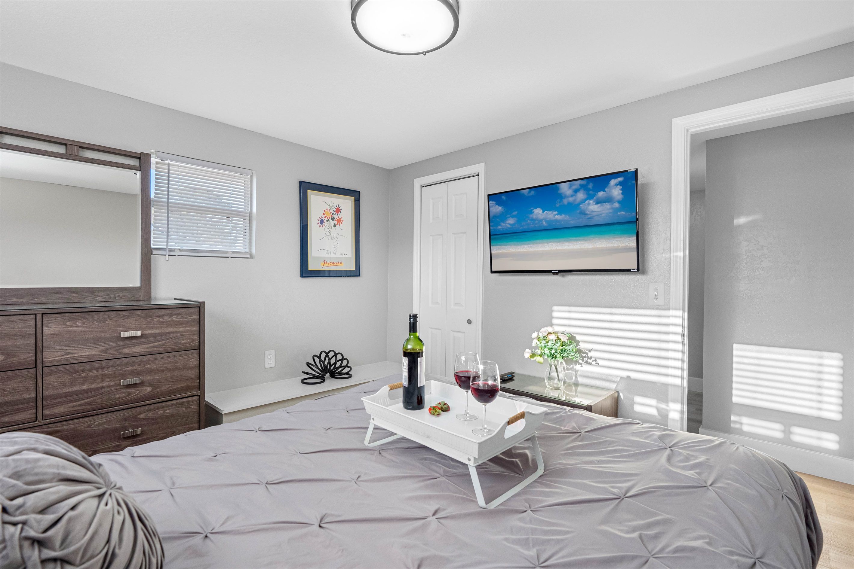 Un dormitorio con cama, cómoda, televisor en la pared y ventana con persianas.