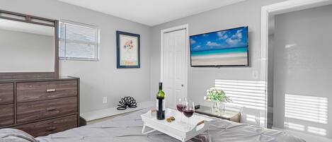 Un dormitorio con cama, cómoda, televisor en la pared y ventana con persianas.