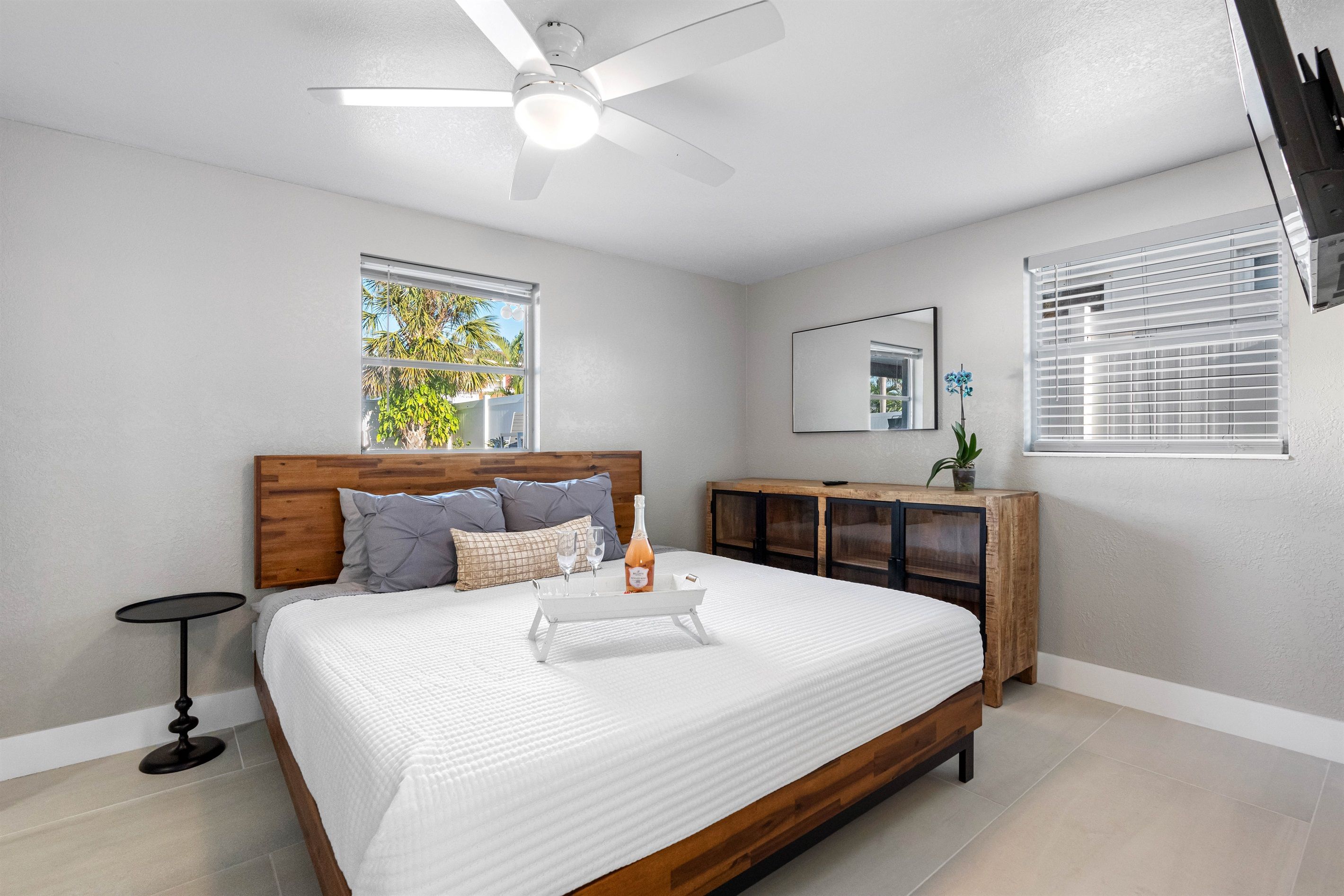 A bedroom with a bed, a ceiling fan, a bedside table, a nightstand, a dresser, and a window with blinds.