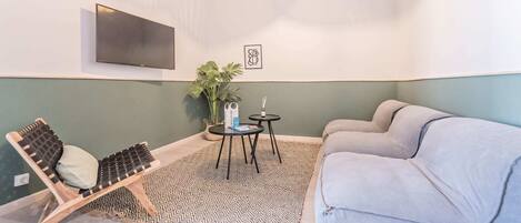 Un salon moderne avec un canapé gris, une chaise en bois, un tapis à motifs, une petite table ronde, une plante en pot et un téléviseur fixé au mur.
