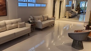 A modern living room with a beige sofa, a coffee table, and a decorative chair. There is a large window with blinds, a ceiling fan, and a Christmas tree. - Santos Linda !! 5 min from the beach ! (SP)