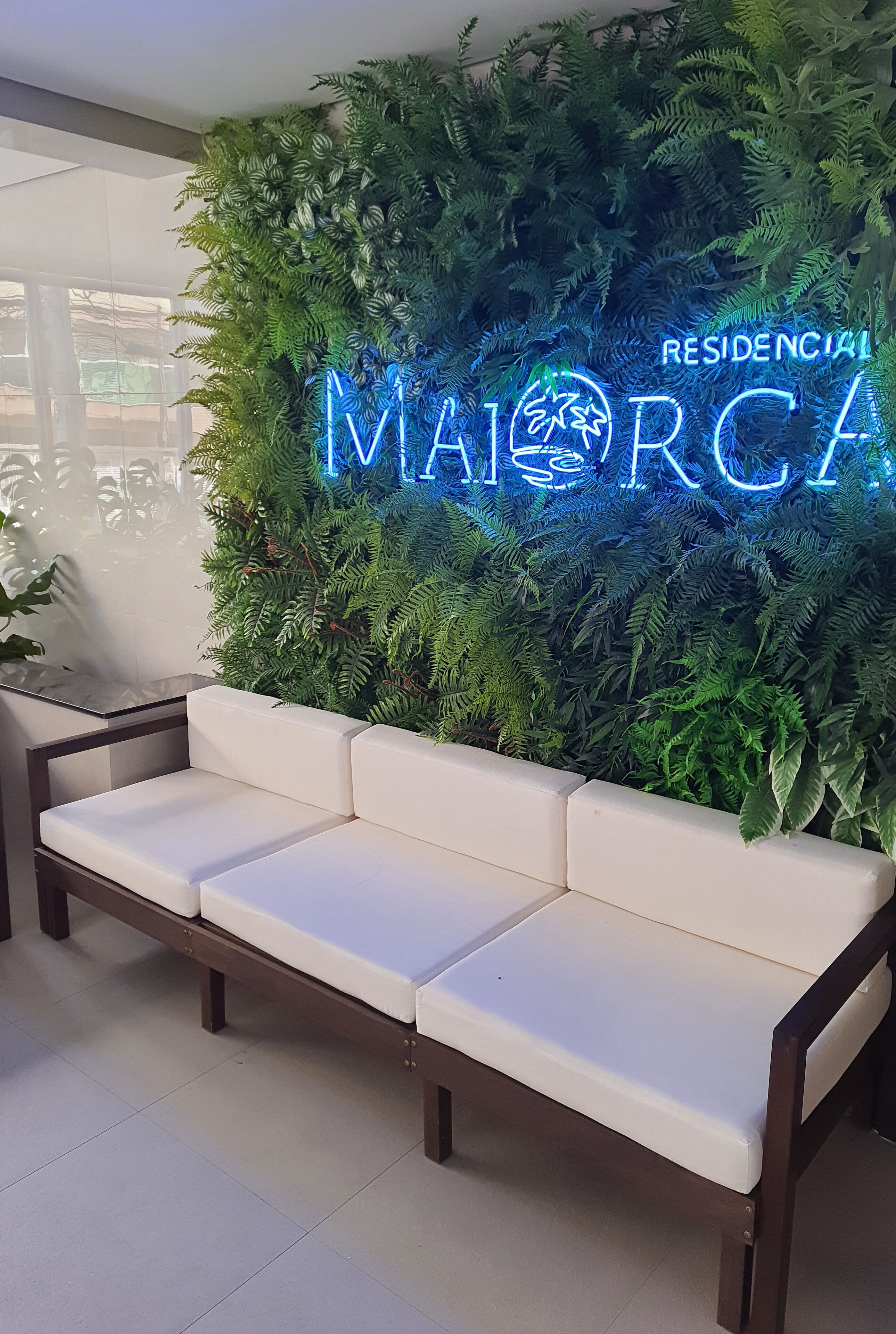 Un área de recepción con un letrero de neón que dice "MAIORCA RESIDENCIAL" y un sofá blanco frente a una pared verde cubierta de plantas.