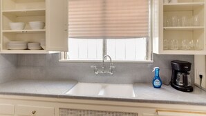 A kitchen with a sink, a window with blinds, and shelves with bowls and glasses.