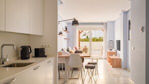 A modern kitchen with a built-in oven, a coffee machine, and a sink. There is a dining area with a table and chairs, and a balcony with plants visible through the glass doors.