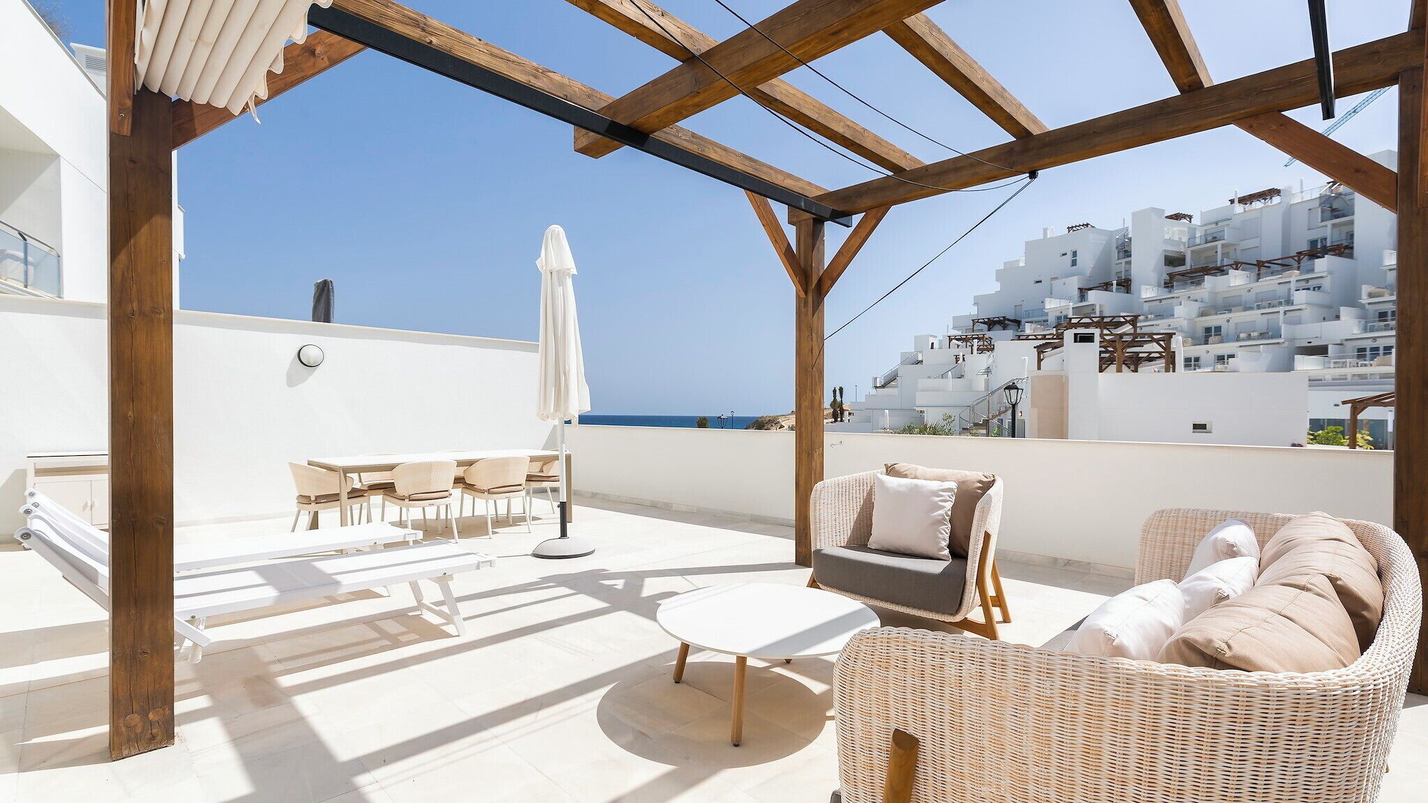 Una terraza en la azotea con una pérgola de madera, mobiliario exterior y vistas a edificios blancos y al mar.