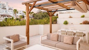 A rooftop terrace with a wooden pergola, white furniture, and a view of the cityscape.