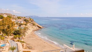 A coastal view with a sandy beach, a clear blue sea, and buildings on a hillside.