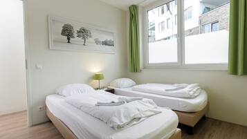 A hotel room with two beds, a nightstand with a lamp, a painting of trees, and a window with green curtains.