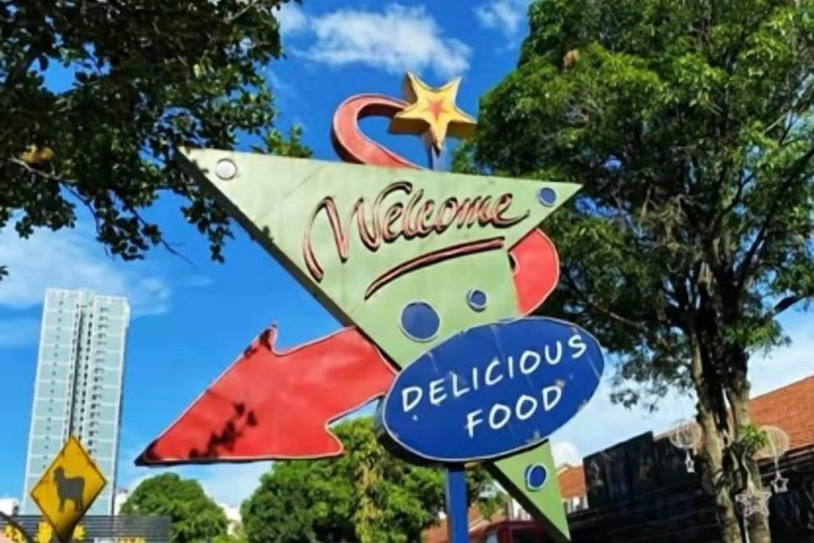 Un cartell de benvinguda colorit amb les paraules 'Welcome' i 'Delicious Food' davant d'un fons de trees i un edifici.