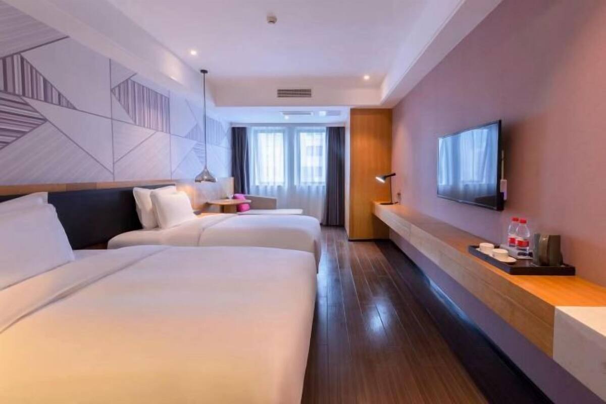 A hotel room with two beds, a TV, a bedside table with a bottle and glasses, and a wall-mounted TV.