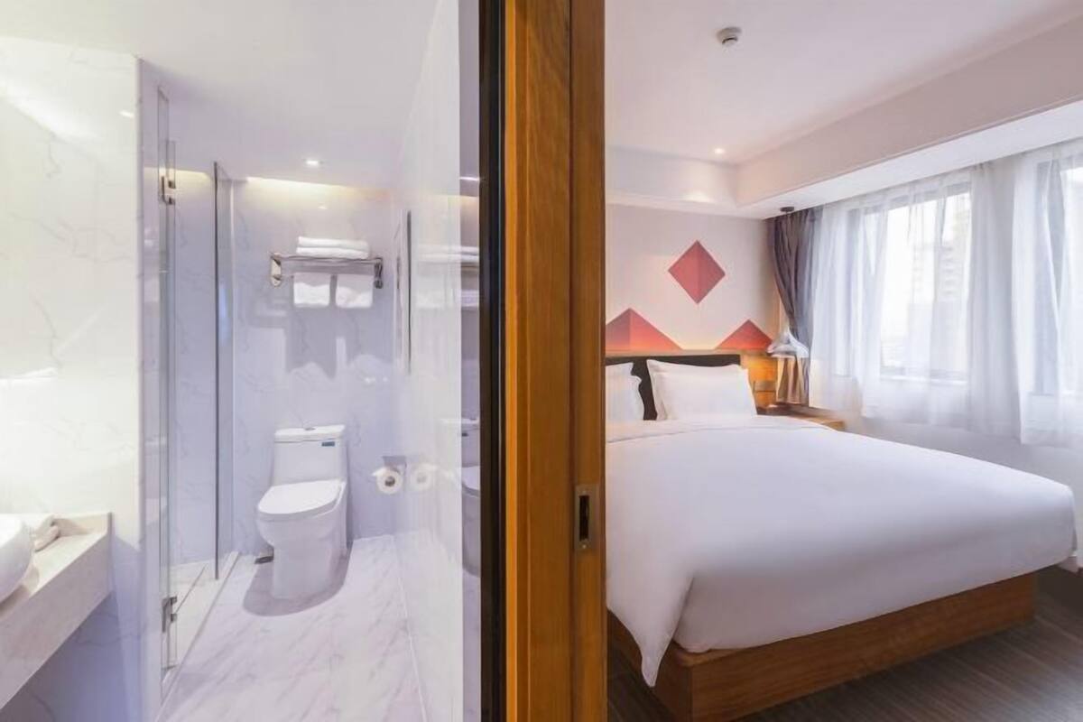 A hotel room with a glass shower enclosure, a bed with a wooden headboard, and a view of the cityscape through the window.