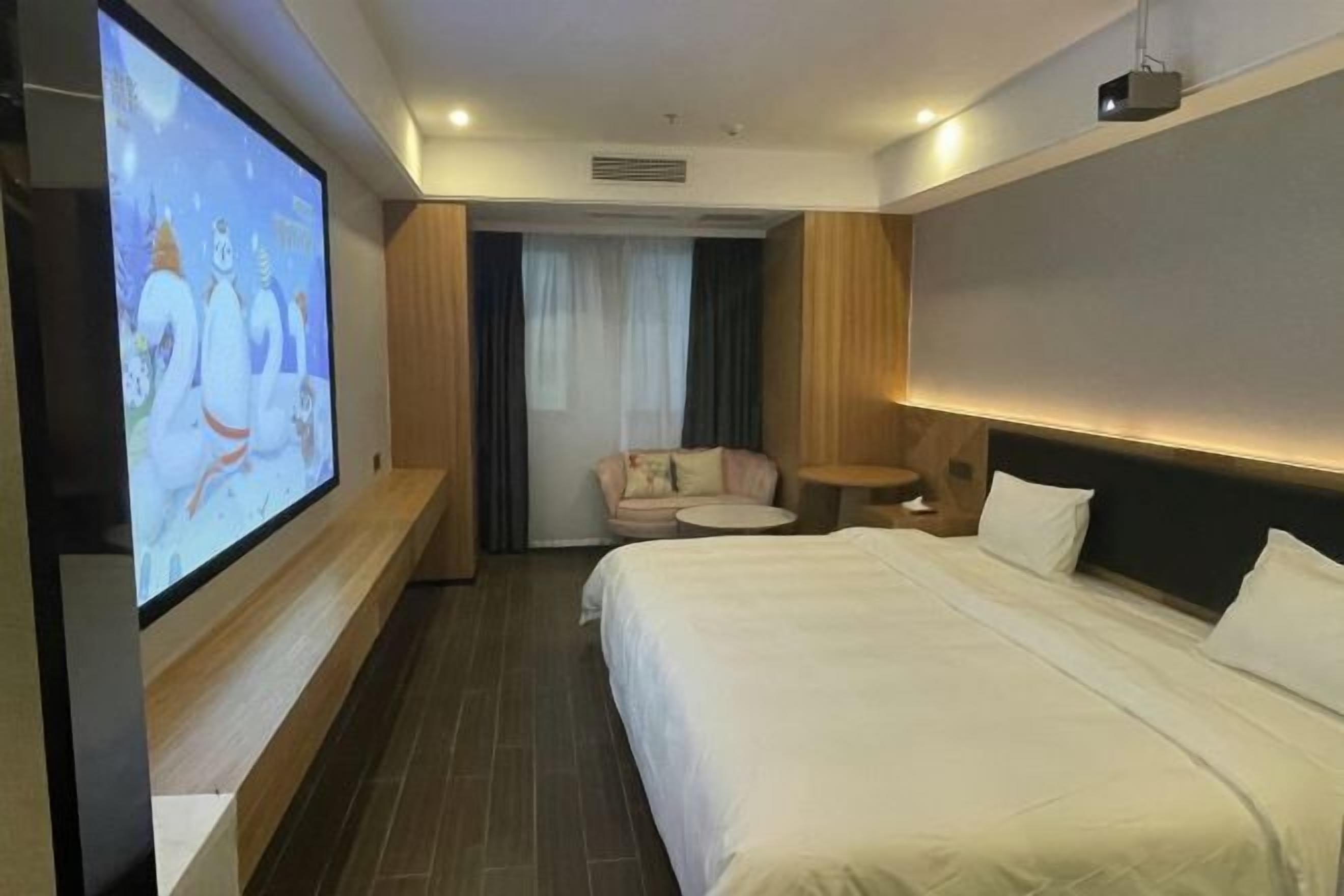 Quarto de hotel com cama grande, televisão fixada na parede, mesa redonda pequena e área de estar com uma poltrona e um sofá.
