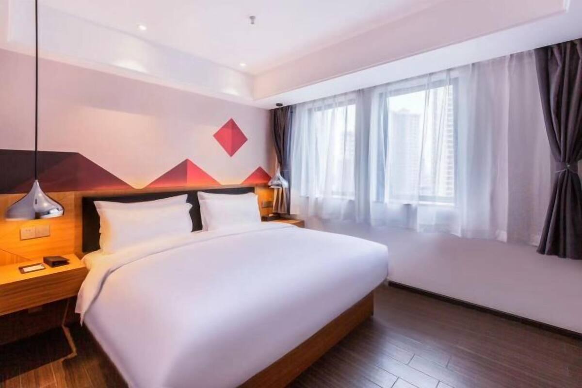 A modern hotel room with a large bed, a nightstand, a lamp, and a window with curtains.