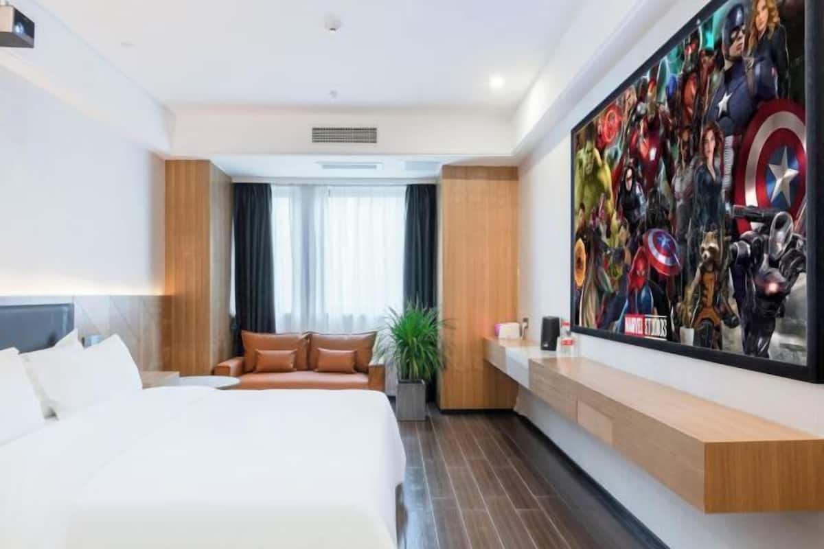 A hotel room with a large bed, a sofa, a desk, and a wall-mounted TV displaying a movie poster.