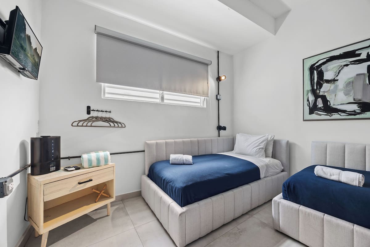 A modern bedroom with two beds, a wooden dresser, a TV, and a painting on the wall.