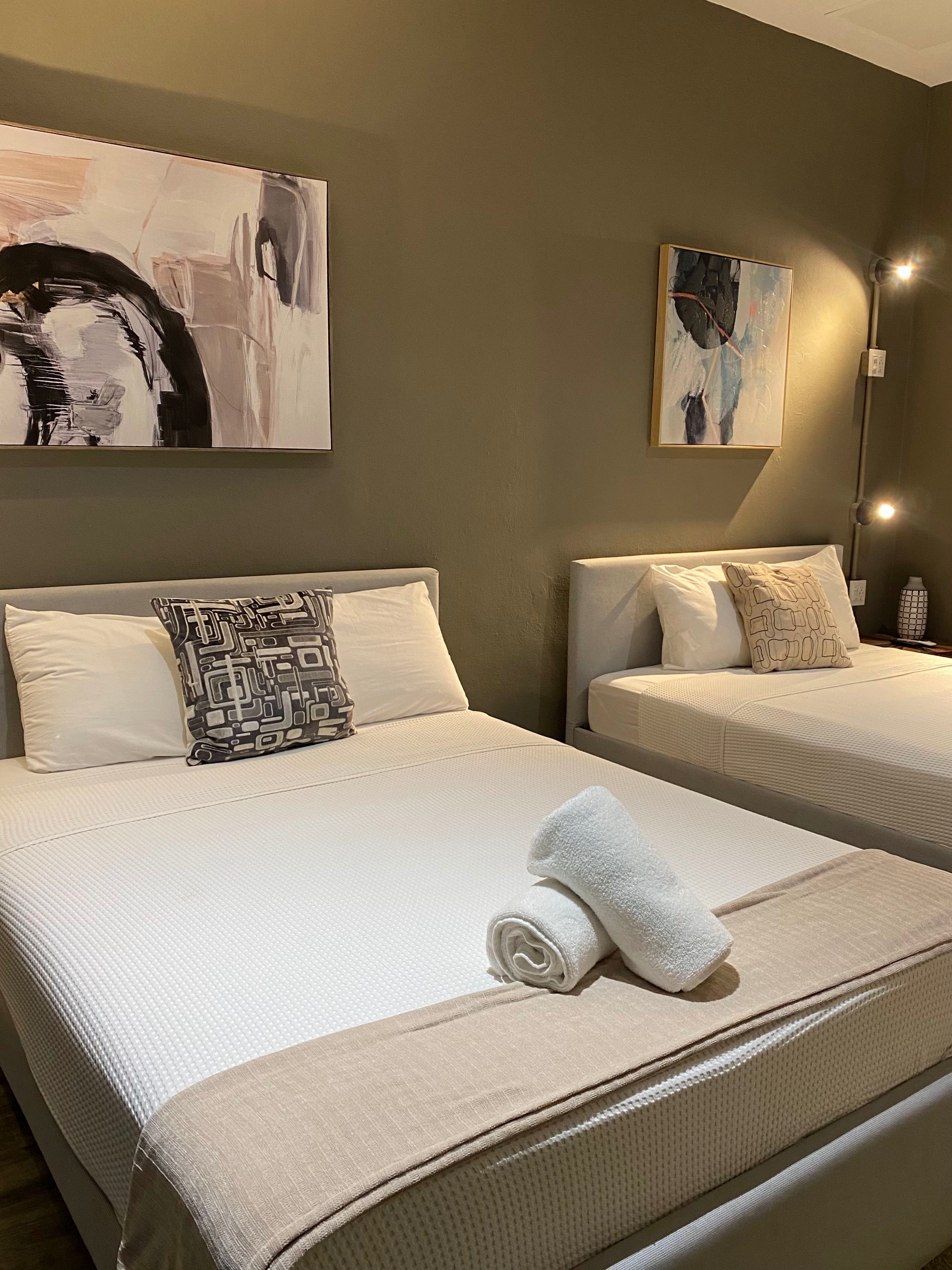 Une chambre d’hôtel avec deux lits, chacun agrémenté de draps et d’oreillers blancs, une serviette roulée posée sur le lit au premier plan, et des œuvres d’art abstraites accrochées aux murs.