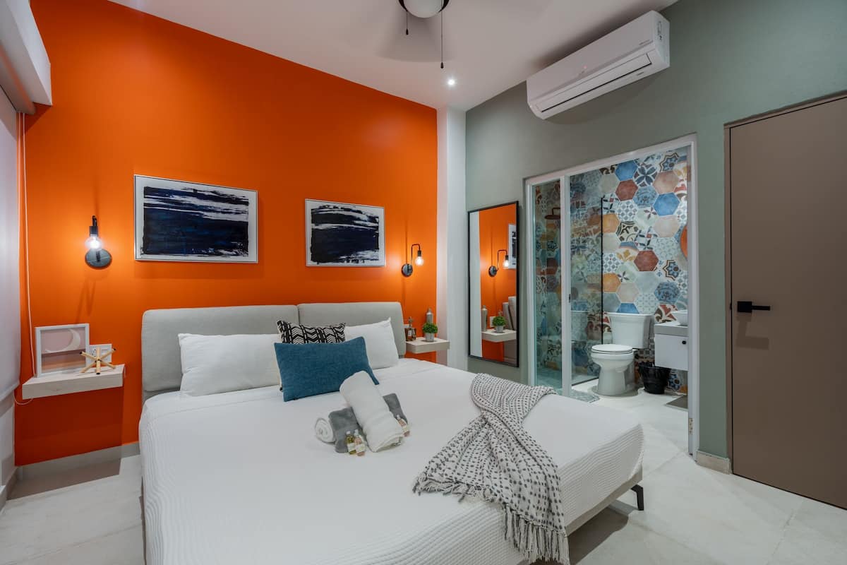 A modern bedroom with a large bed, orange walls, and a bathroom with a tiled shower and toilet.