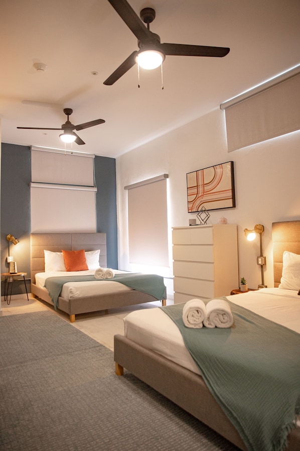 A modern hotel room with two beds, a ceiling fan, a large window, and a wall-mounted artwork. - La Buena Vida Inn (San Juan)