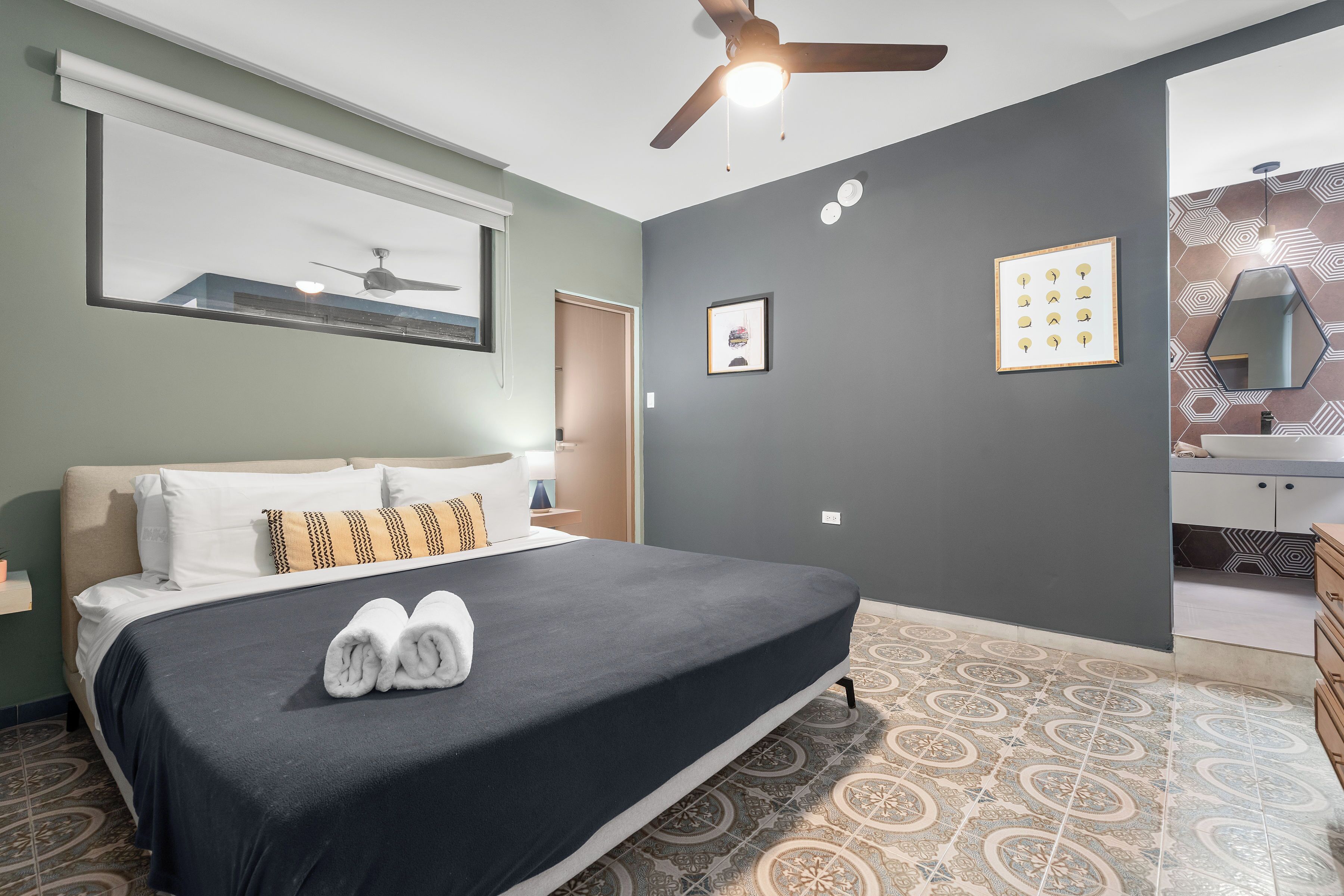 Ein Hotelzimmer mit Bett, Deckenventilator, Fenster und einem durchgegangenen Tür sichtbaren Badezimmer.