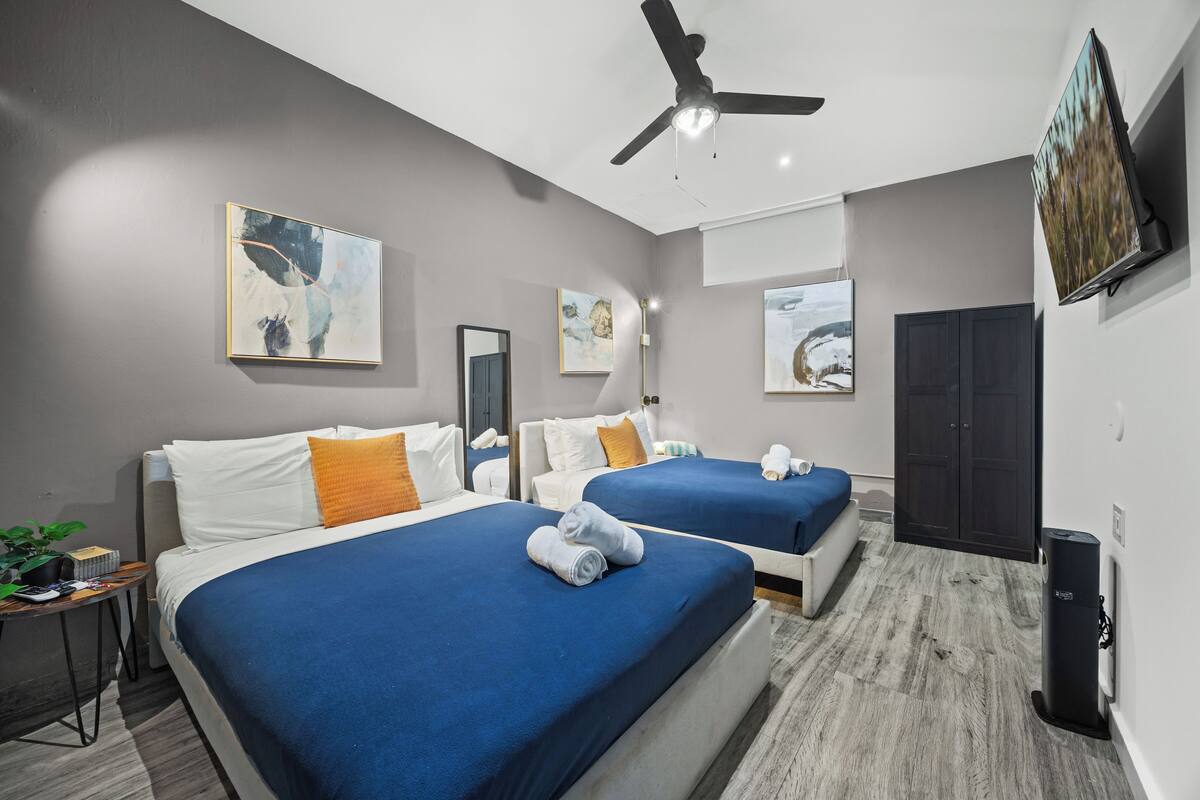 A hotel room with two beds, a ceiling fan, artwork on the walls, and a TV mounted on the wall.