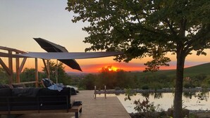 Terraza de madera con sofá y sombrilla, con vista a un cuerpo de agua y al atardecer.