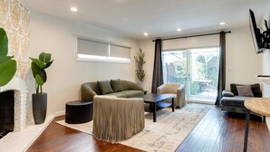 Una sala de estar moderna con sofá, sillón y mesa de centro. Hay una chimenea, ventanas amplias y un televisor montado en la pared.