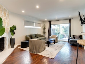 Ein modernes Wohnzimmer mit Sofa, Sessel und Couchtisch. Ein Kamin, große Fenster und ein an der Wand befestigter Fernseher sind ebenfalls vorhanden.