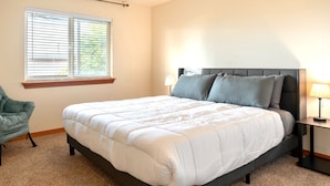 A bedroom with a large bed, a window with blinds, a chair, and a nightstand with a lamp. - Dune Breeze - Wifi and Hot tub (Pacific Beach)