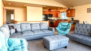 A spacious living room with a grey sectional sofa, a teal armchair, and a grey ottoman. A kitchen with wooden cabinets and stainless steel appliances is visible in the background. - Dune Breeze - Wifi and Hot tub (Pacific Beach)