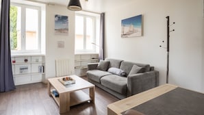 A modern living room with a grey sofa, wooden coffee table, and a view of the outdoors through large windows. - Le P'tit Cancalais - Apt à 300 m de la mer (Cancale)