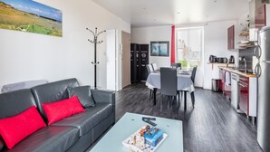 A modern apartment with a living room, dining area, and kitchen. It features a black leather sofa, a dining table with chairs, a kitchen with a refrigerator and stove, and a coat rack.