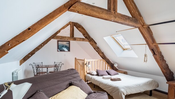 A cozy attic bedroom with exposed wooden beams, a loft bed, a sofa, and a small dining table.