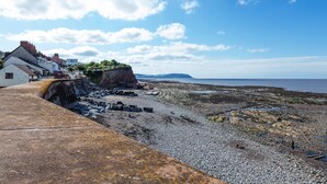 A coastal area with a rocky beach, a cliff with buildings, and a clear sky. - Sea Spray (Watchet)