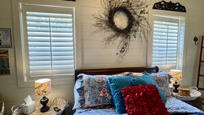 A bedroom with a bed, two bedside tables, a lamp, a basket, a tissue box, a decorative wreath, and a window with blinds.