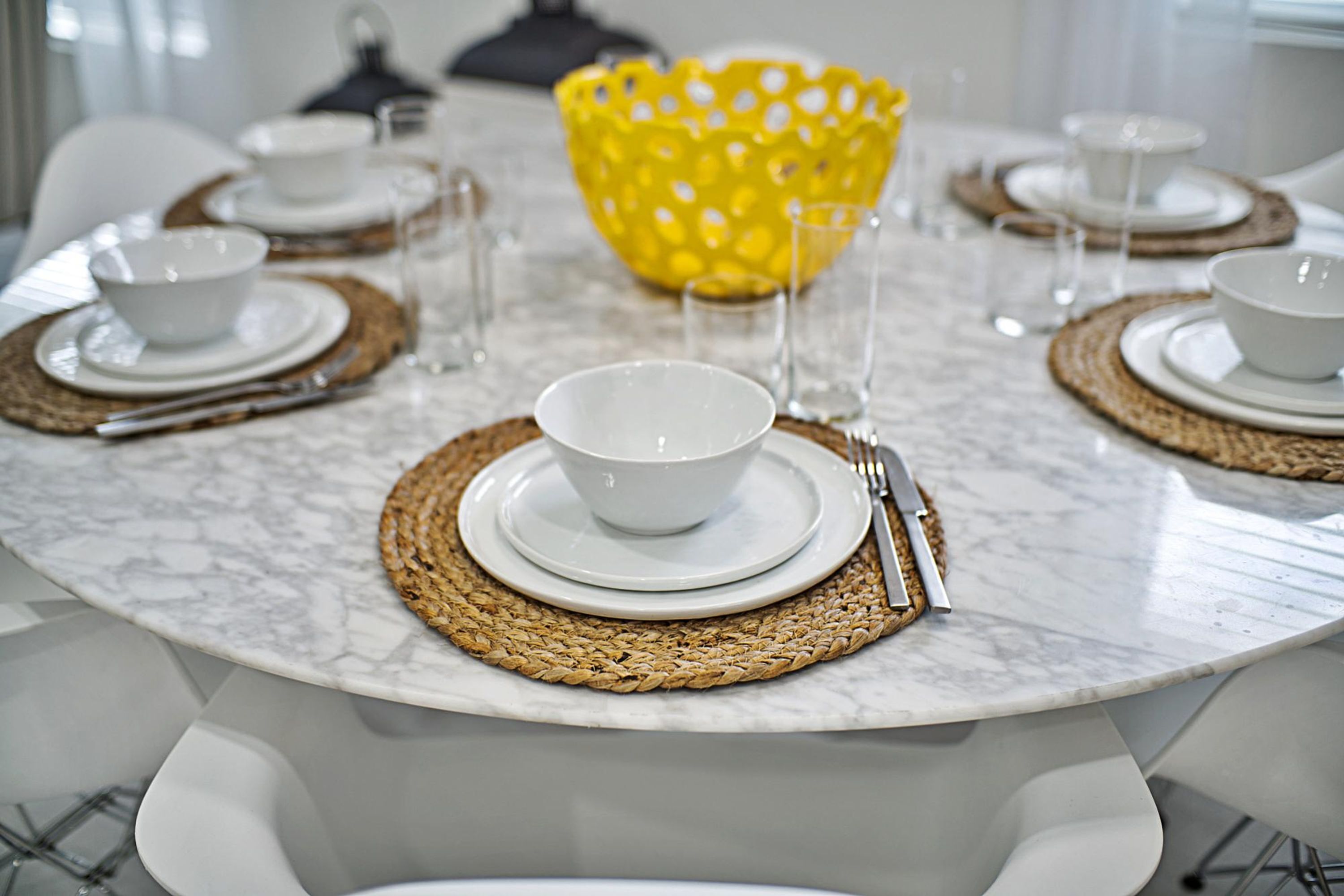 A round table set with white plates, bowls, and cutlery, accompanied by woven placemats and clear glasses.
